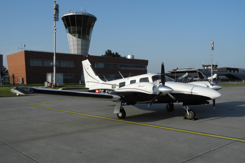 Menší dvoumotorové letadlo Piper Seneca na odbavovací ploše před řídicí věží a budovou letiště Brno-Tuřany