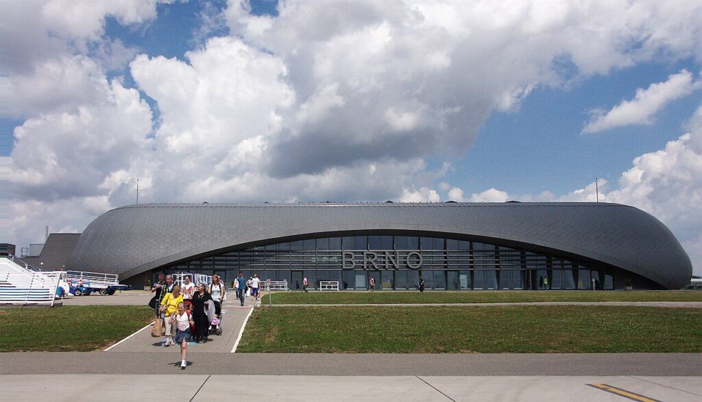 Prosklená fasáda terminálu Letiště Brno-Tuřany s nápisem BRNO a skupinou cestujících s kufry na chodníku před budovou